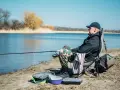 Mężczyzna siedzi na krześle nad jeziorem, stosując metody łowienia ryb. Wędka zarzucona, obok torba i pudełko na przynęty.