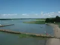Letnie popołudnie nad jeziorem. Ludzie kąpią się, opalają na plaży i spacerują po drewnianym molo. W oddali widać żaglówki. Wypływa z jeziora roś.