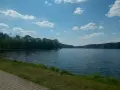Spokojne jezioro soczewka otoczone zielonym lasem, z piaszczystą ścieżką i trawą na pierwszym planie.