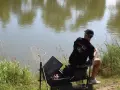 Wędkarz siedzi nad wodą, przygotowany do zawodów feederowych. Obok niego podbierak i skrzynka z akcesoriami.