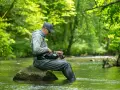 Mężczyzna siedzi na kamieniu w rzece, przygotowując wędkę. Pokazuje, jak łowić na rzece w otoczeniu zieleni.