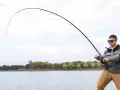 Mężczyzna w okularach przeciwsłonecznych łowi ryby nad jeziorem. Wędka wygięta w łuk, widać napięcie żyłki. Street fishing w miejskim otoczeniu.