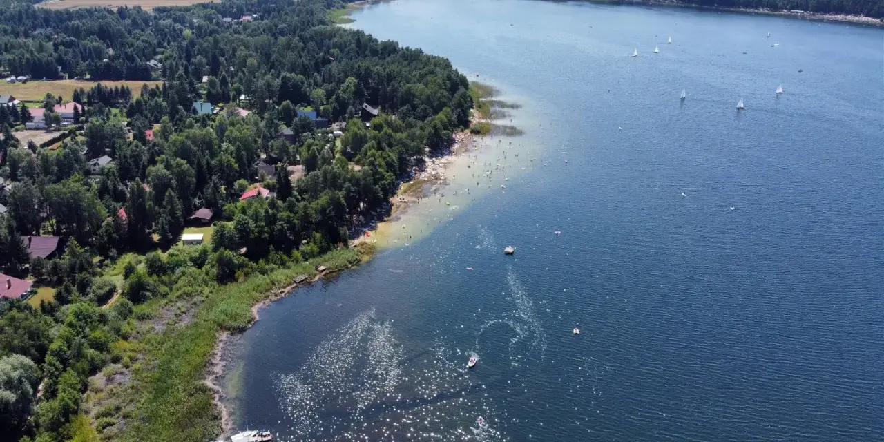 Malownicze jeziora w pomorskim, z piaszczystą plażą, lasem i domkami letniskowymi. Na wodzie żaglówki i kąpielowicze.