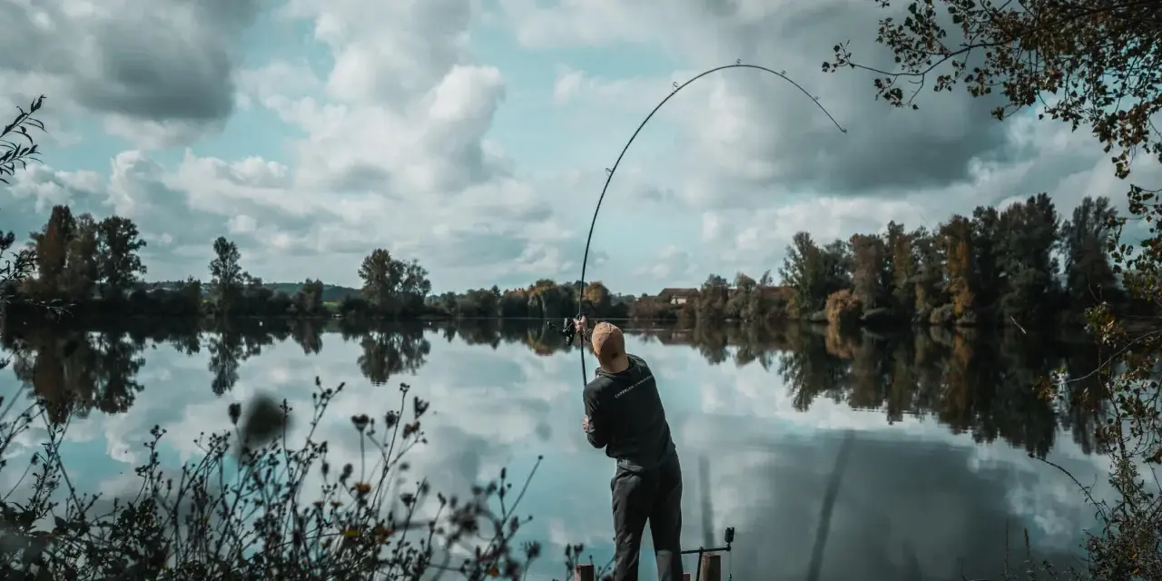 Wędkarz zgiętą wędką walczy z rybą na spokojnej rzece. Na brzegu widać drewniany pomost i roślinność.
