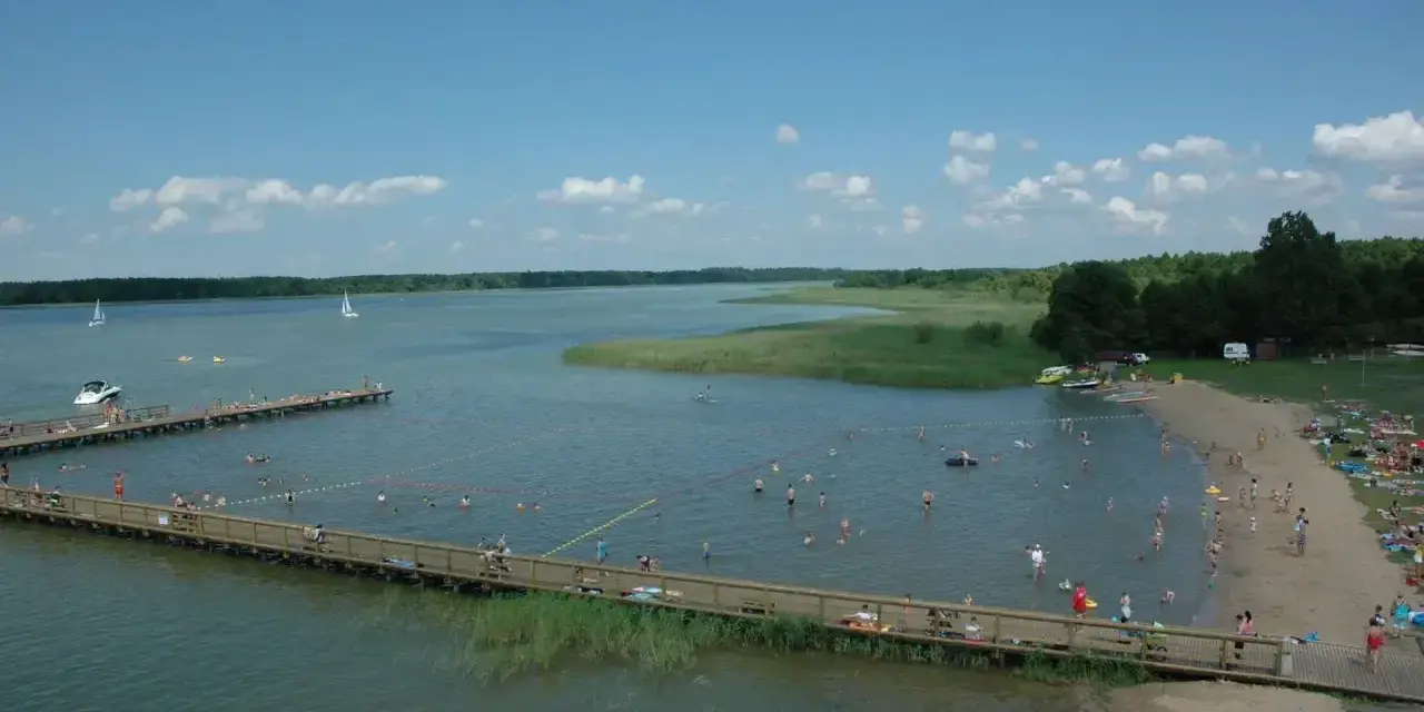 Letnie popołudnie nad jeziorem. Ludzie kąpią się, opalają na plaży i spacerują po drewnianym molo. W oddali widać żaglówki. Wypływa z jeziora roś.
