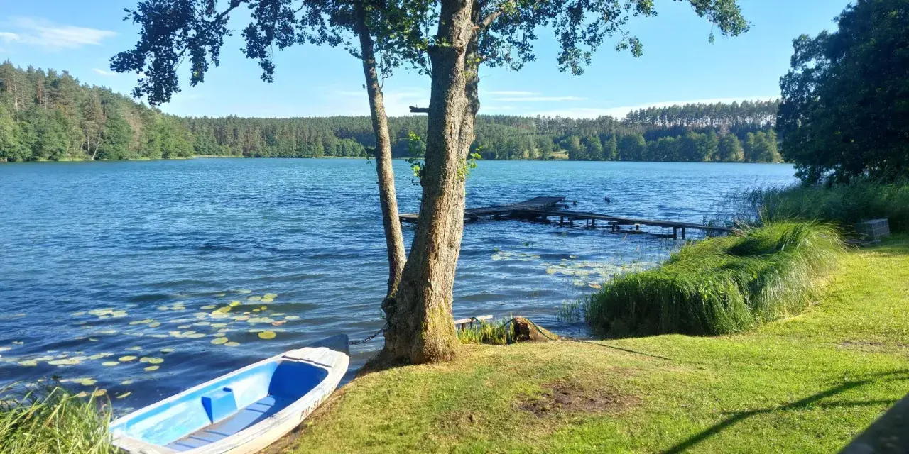 Spokojne jezioro zamkowe z łódką przy brzegu, drewnianym pomostem i lasem w tle.
