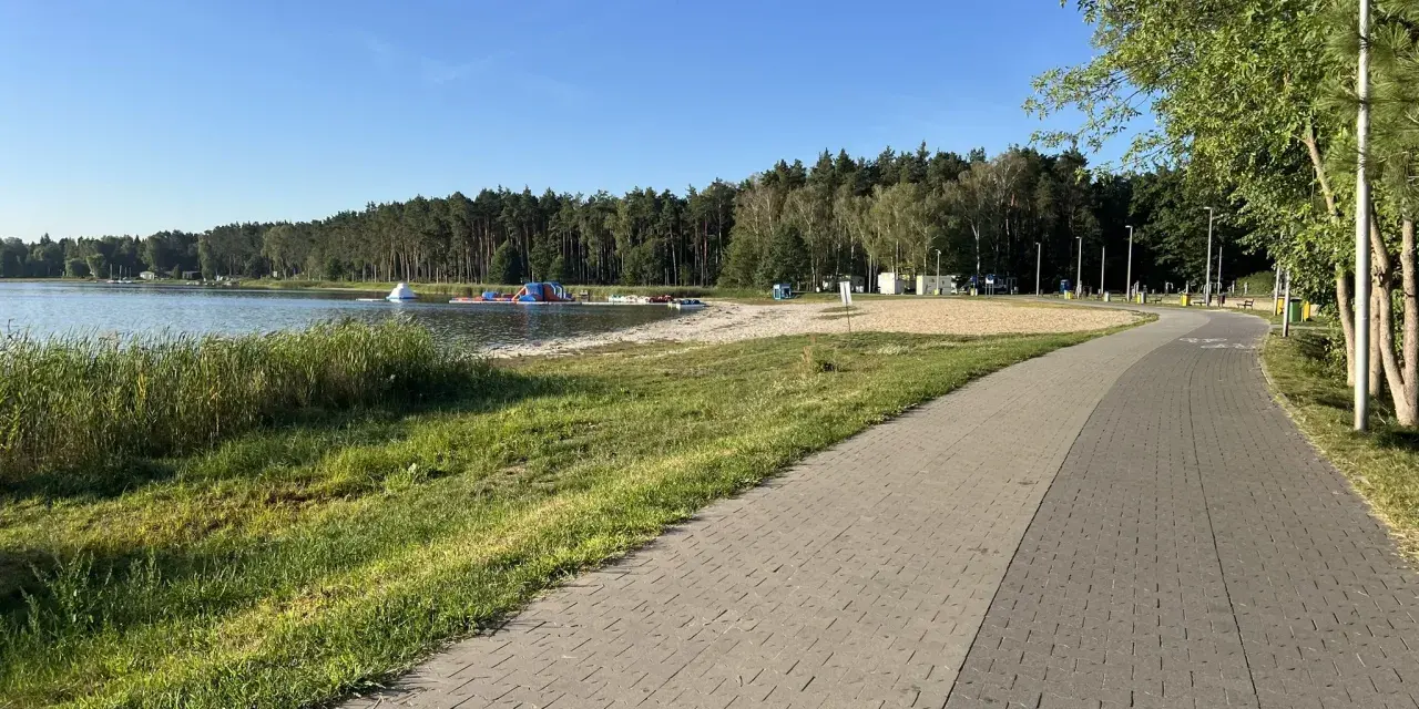 Słoneczny dzień nad jeziorem Firlejowskim. Woda, piaszczysta plaża z dmuchanymi atrakcjami i ścieżka rowerowa.