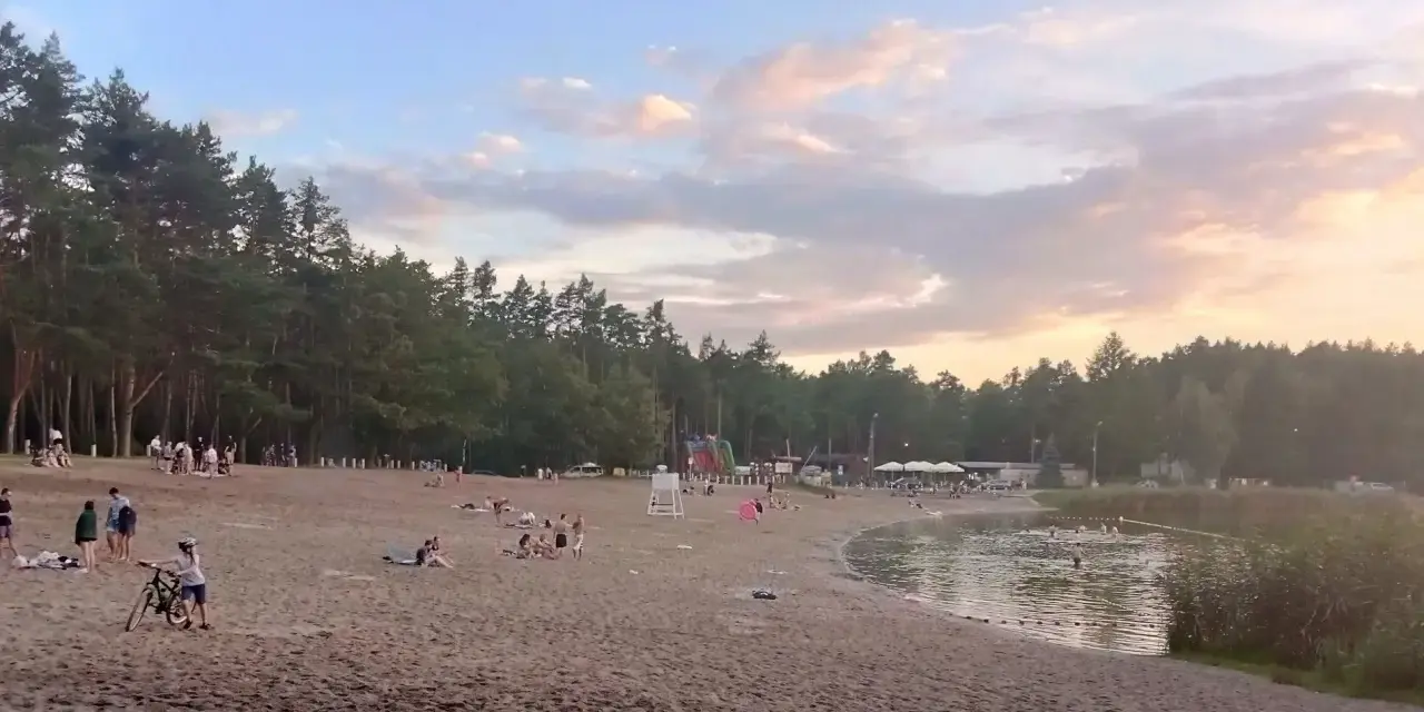Plaża nad jeziorem Jelcz Laskowice tętni życiem. Ludzie odpoczywają na piasku, kąpią się w wodzie, a dzieci bawią się na dmuchanych zjeżdżalniach.