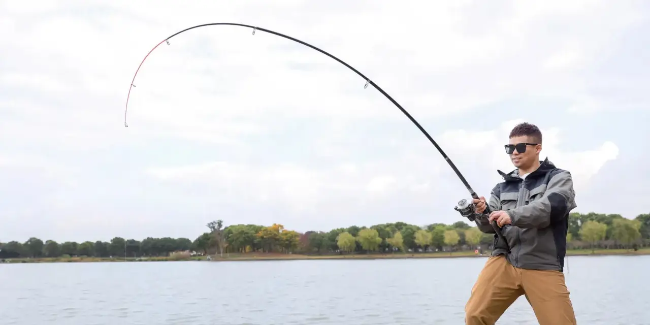 Mężczyzna w okularach przeciwsłonecznych łowi ryby nad jeziorem. Wędka wygięta w łuk, widać napięcie żyłki. Street fishing w miejskim otoczeniu.