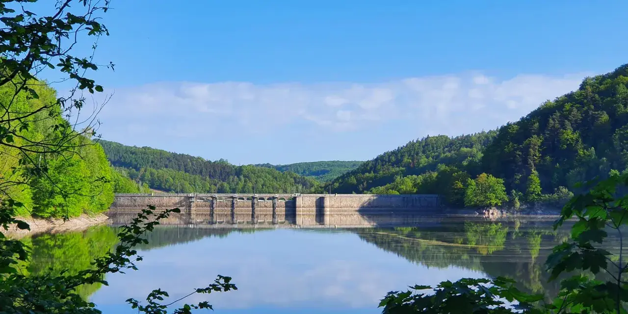 Spokojne, zielone jezioro odbija zalesione wzgórza i kamienną tamę.