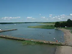 Letnie popołudnie nad jeziorem. Ludzie kąpią się, opalają na plaży i spacerują po drewnianym molo. W oddali widać żaglówki. Wypływa z jeziora roś.