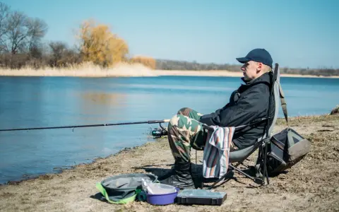 Mężczyzna siedzi na krześle nad jeziorem, stosując metody łowienia ryb. Wędka zarzucona, obok torba i pudełko na przynęty.