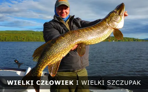 Szczęśliwy wędkarz trzyma ogromnego szczupaka na tle jeziora i zielonych łąk.
