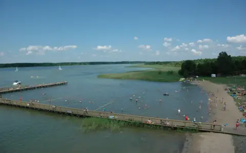 Letnie popołudnie nad jeziorem. Ludzie kąpią się, opalają na plaży i spacerują po drewnianym molo. W oddali widać żaglówki. Wypływa z jeziora roś.