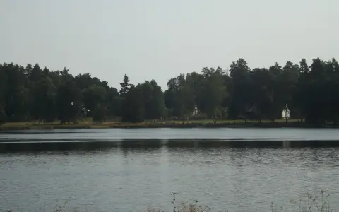 Spokojne jezioro zmarłe otoczone gęstym lasem. Na brzegu widać kilka domków.