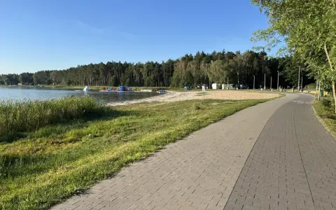 Słoneczny dzień nad jeziorem Firlejowskim. Woda, piaszczysta plaża z dmuchanymi atrakcjami i ścieżka rowerowa.