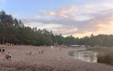Plaża nad jeziorem Jelcz Laskowice tętni życiem. Ludzie odpoczywają na piasku, kąpią się w wodzie, a dzieci bawią się na dmuchanych zjeżdżalniach.
