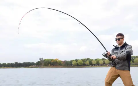 Mężczyzna w okularach przeciwsłonecznych łowi ryby nad jeziorem. Wędka wygięta w łuk, widać napięcie żyłki. Street fishing w miejskim otoczeniu.