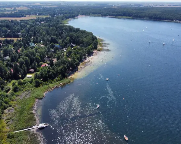 Malownicze jeziora w pomorskim, z piaszczystą plażą, lasem i domkami letniskowymi. Na wodzie żaglówki i kąpielowicze.