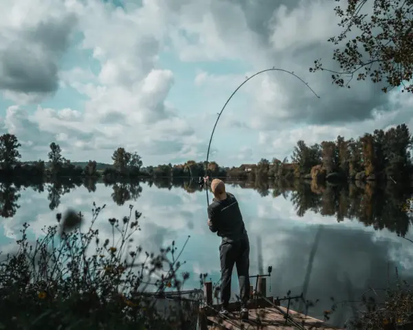 Wędkarz zgiętą wędką walczy z rybą na spokojnej rzece. Na brzegu widać drewniany pomost i roślinność.
