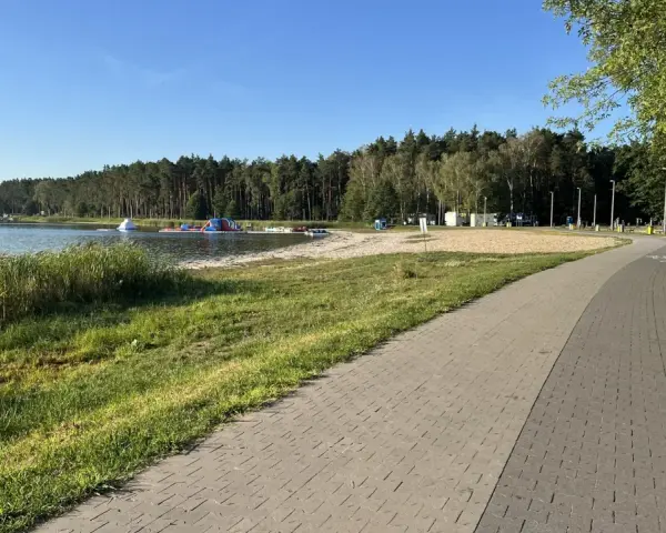Słoneczny dzień nad jeziorem Firlejowskim. Woda, piaszczysta plaża z dmuchanymi atrakcjami i ścieżka rowerowa.
