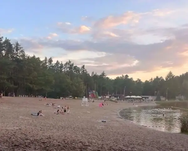 Plaża nad jeziorem Jelcz Laskowice tętni życiem. Ludzie odpoczywają na piasku, kąpią się w wodzie, a dzieci bawią się na dmuchanych zjeżdżalniach.
