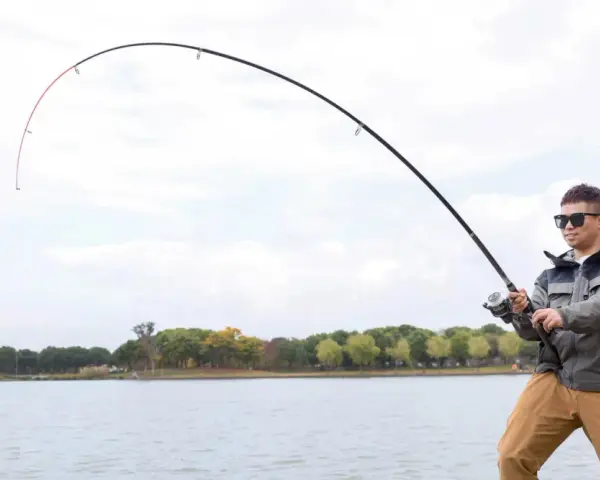 Mężczyzna w okularach przeciwsłonecznych łowi ryby nad jeziorem. Wędka wygięta w łuk, widać napięcie żyłki. Street fishing w miejskim otoczeniu.