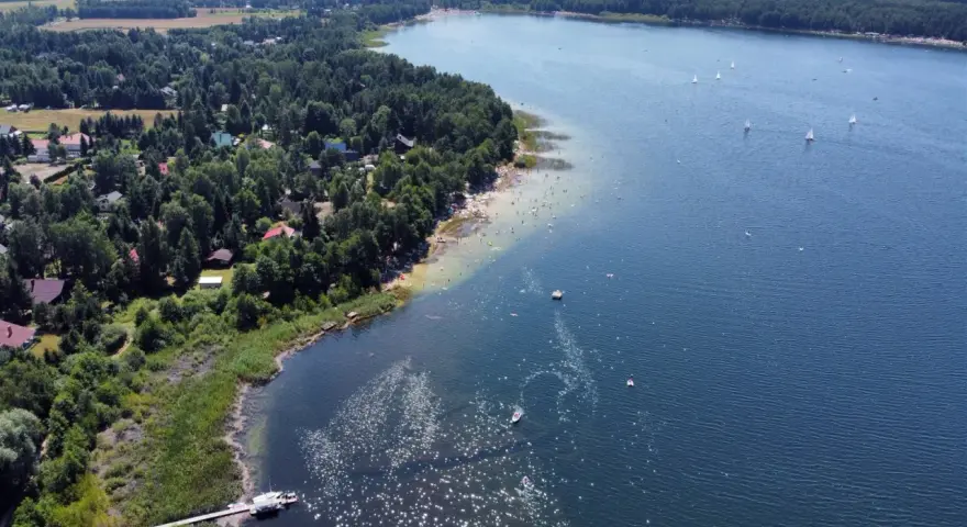 Malownicze jeziora w pomorskim, z piaszczystą plażą, lasem i domkami letniskowymi. Na wodzie żaglówki i kąpielowicze.