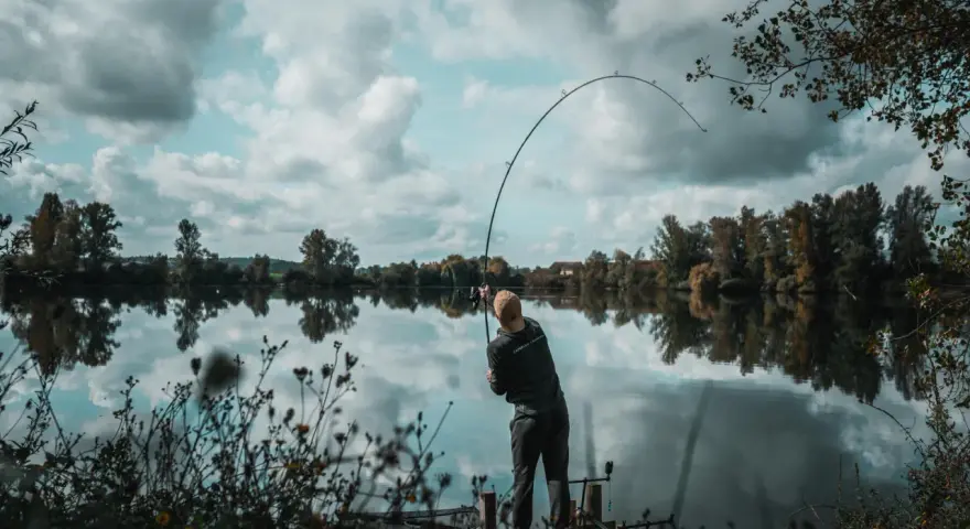 Wędkarz zgiętą wędką walczy z rybą na spokojnej rzece. Na brzegu widać drewniany pomost i roślinność.
