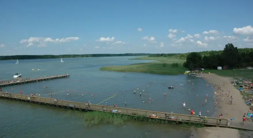 Letnie popołudnie nad jeziorem. Ludzie kąpią się, opalają na plaży i spacerują po drewnianym molo. W oddali widać żaglówki. Wypływa z jeziora roś.