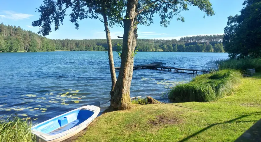 Spokojne jezioro zamkowe z łódką przy brzegu, drewnianym pomostem i lasem w tle.