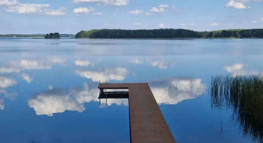 Drewniane molo prowadzi do spokojnego jeziora nisko, gdzie błękitne niebo z białymi chmurami odbija się w tafli wody.