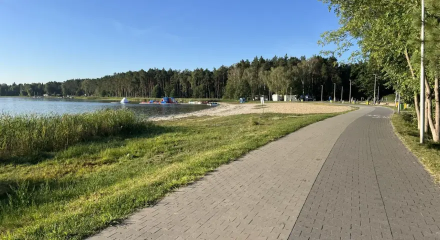 Słoneczny dzień nad jeziorem Firlejowskim. Woda, piaszczysta plaża z dmuchanymi atrakcjami i ścieżka rowerowa.
