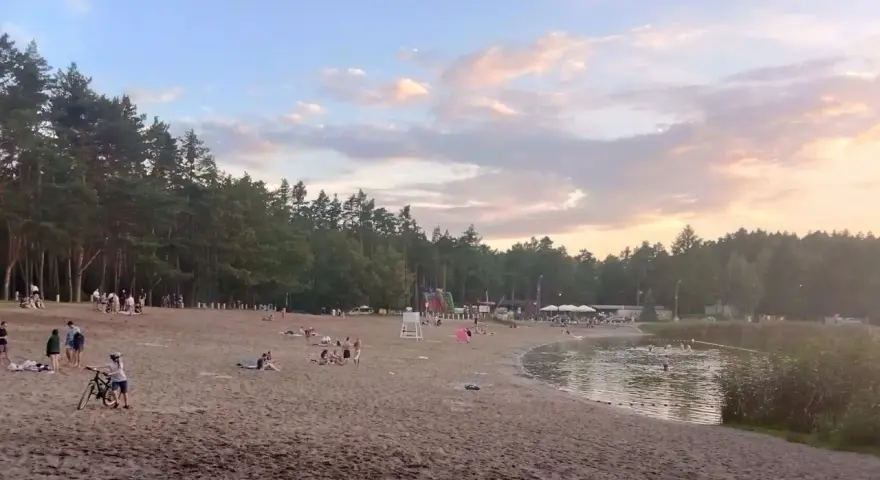 Plaża nad jeziorem Jelcz Laskowice tętni życiem. Ludzie odpoczywają na piasku, kąpią się w wodzie, a dzieci bawią się na dmuchanych zjeżdżalniach.