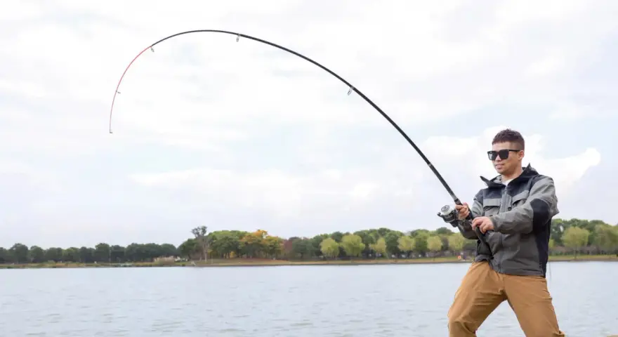 Mężczyzna w okularach przeciwsłonecznych łowi ryby nad jeziorem. Wędka wygięta w łuk, widać napięcie żyłki. Street fishing w miejskim otoczeniu.
