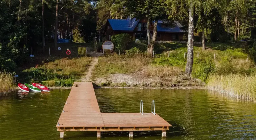 Drewniany pomost nad jeziorem Zagnanie, obok trzy kajaki. W tle dom wśród drzew.