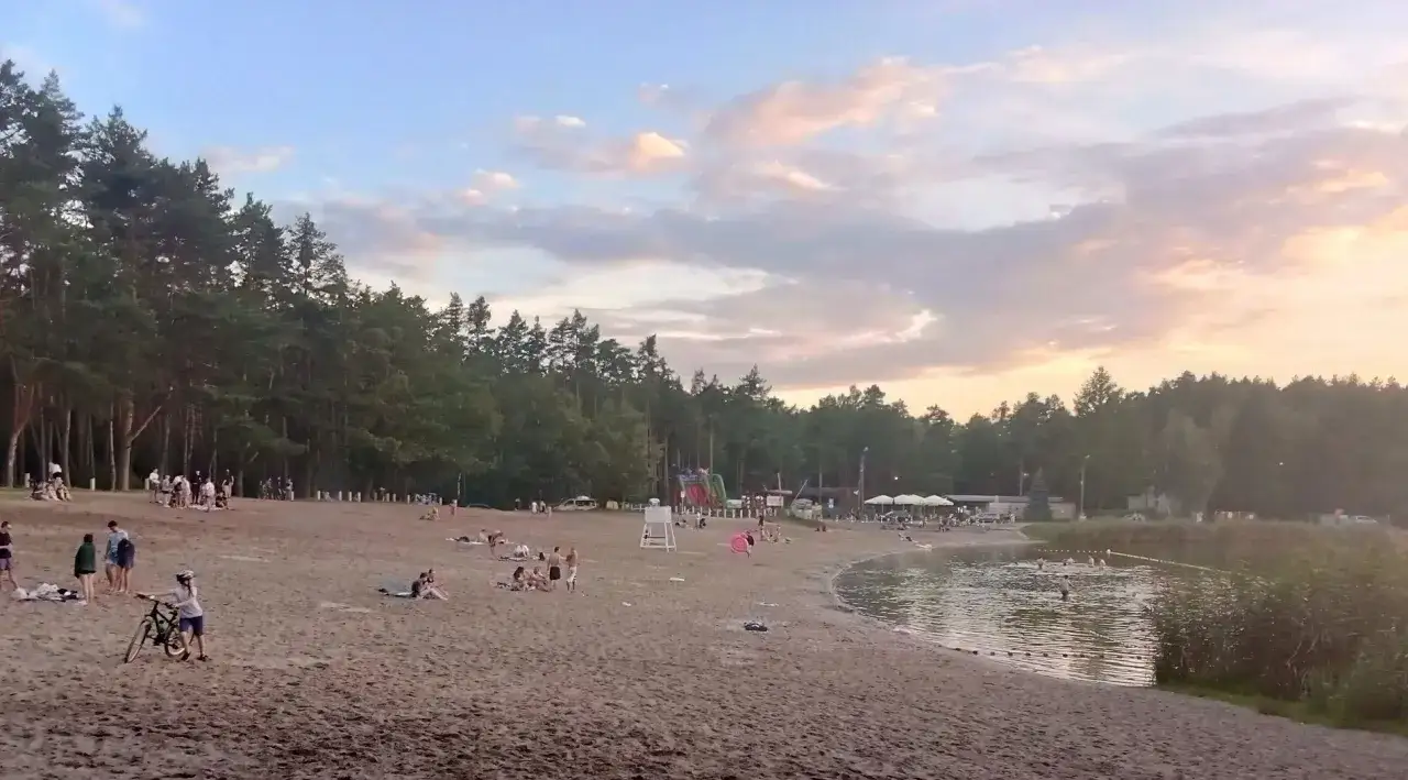 Plaża nad jeziorem Jelcz Laskowice tętni życiem. Ludzie odpoczywają na piasku, kąpią się w wodzie, a dzieci bawią się na dmuchanych zjeżdżalniach.