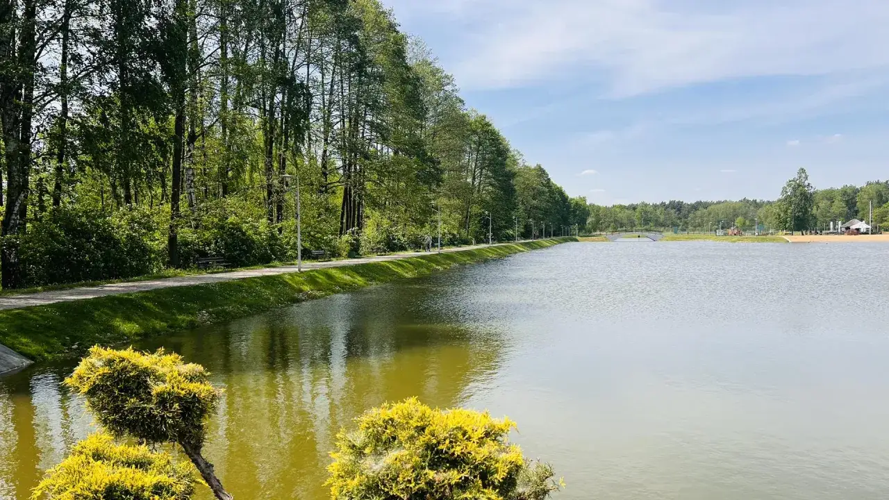 Malownicze jezioro Kunów otoczone zielenią, z alejką spacerową i mostkiem w oddali. Idealne miejsce na relaks.
